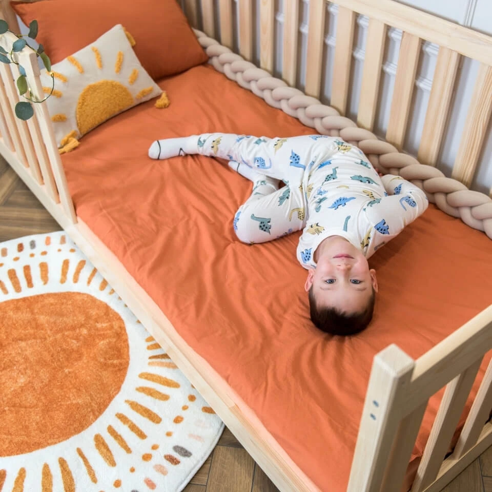Chlapec leží v borovicové nízké posteli s vysokými zábranami, dominuje oranžová barva a boho dekorace s motivem slunce.